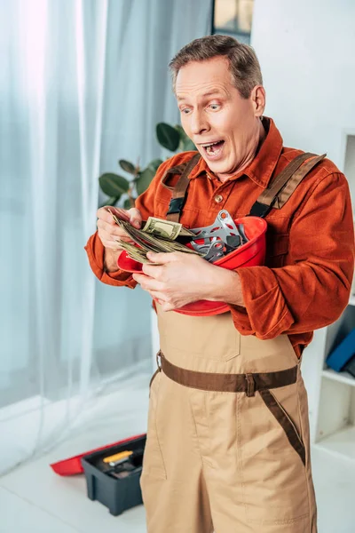 Surprised repairman holding hardhat with money in office — Stock Photo