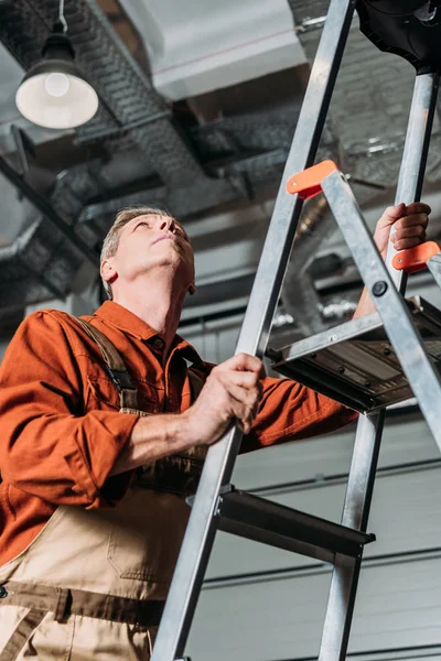 Riparatore in uniforme arancione in piedi su scala in garage — Foto stock