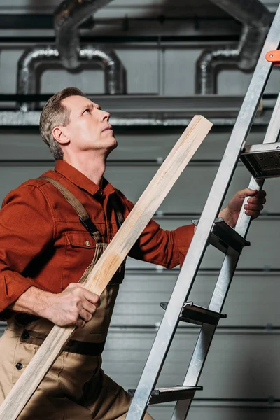 Reparador en uniforme naranja escalando con tablero de madera en mano en escalera en garaje - foto de stock