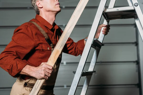 Vista recortada de reparador en uniforme naranja escalando con tablero de madera en mano en escalera en garaje - foto de stock