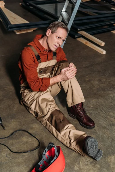 Ремонтник, сидящий на полу и держащий травмированное колено, окружающее оборудование в гараже — стоковое фото