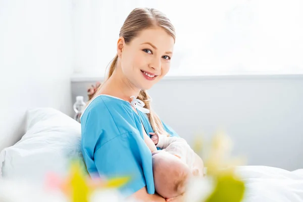 Вибірковий фокус щасливої молодої матері грудного вигодовування новонародженої дитини і посмішка на камеру в лікарняній кімнаті — стокове фото
