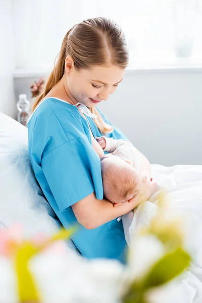 Seitenansicht einer lächelnden jungen Mutter, die ihr Neugeborenes auf dem Bett im Krankenhauszimmer stillt — Stockfoto