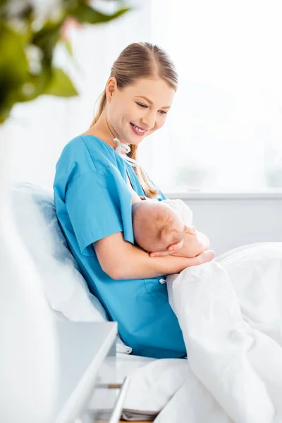 Side view of beautiful happy young mother breastfeeding newborn baby on bed in hospital room — Stock Photo