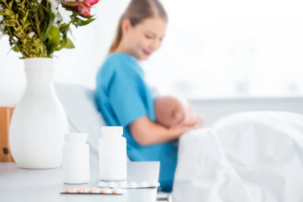 Крупный план таблетки, цветы в вазе и молодая мать кормящая младенца грудью в больничной палате — стоковое фото