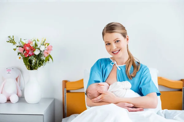 Feliz joven madre sentada en la cama y sonriendo a la cámara mientras amamanta al bebé recién nacido - foto de stock