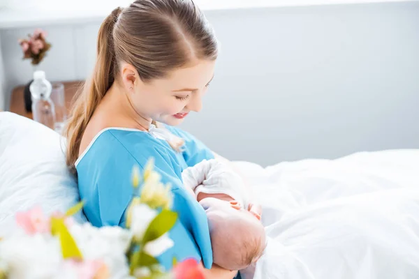 Side view of happy young mother breastfeeding newborn baby on bed in hospital room — Stock Photo