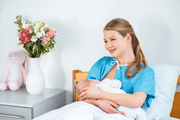 Happy young mother breastfeeding newborn baby and looking away in hospital room — Stock Photo