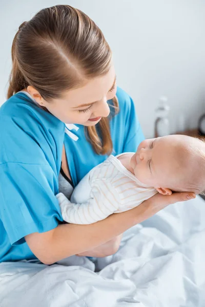 Vista de ángulo alto de la madre joven sentada en la cama del hospital y llevando al bebé adorable - foto de stock