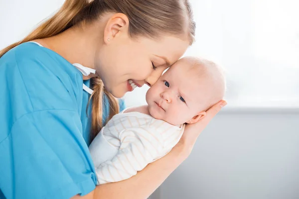 Vista lateral de feliz madre joven llevando y abrazando adorable bebé mirando a la cámara en la habitación del hospital - foto de stock