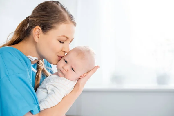 Vista laterale di felice giovane madre che porta e bacia adorabile bambino in camera d'ospedale — Foto stock