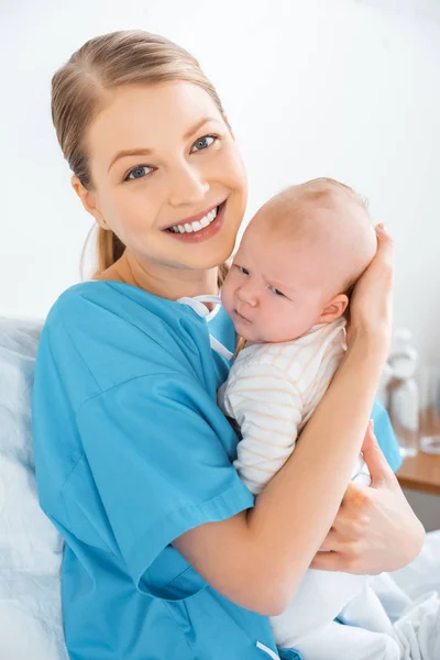 Happy young mother sitting on bed with adorable baby and smiling at camera in hospital room — Stock Photo