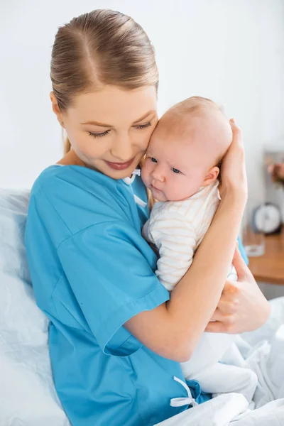 Feliz madre joven sentada en la cama del hospital y abrazando al adorable bebé - foto de stock
