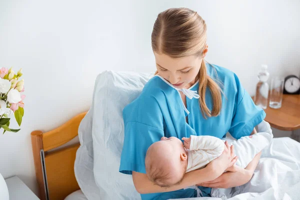 Visão de alto ângulo da jovem mãe sentada na cama do hospital e segurando adorável bebê recém-nascido — Fotografia de Stock