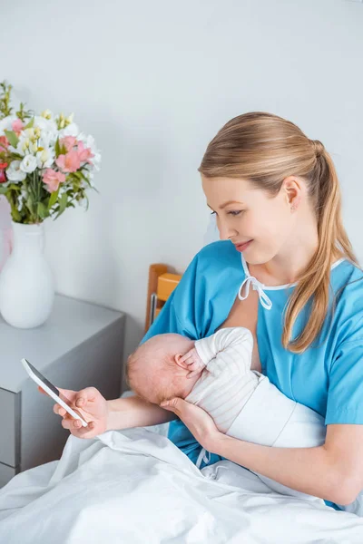 Vista de ángulo alto de la sonriente madre joven amamantando al bebé y utilizando el teléfono inteligente en la habitación del hospital - foto de stock