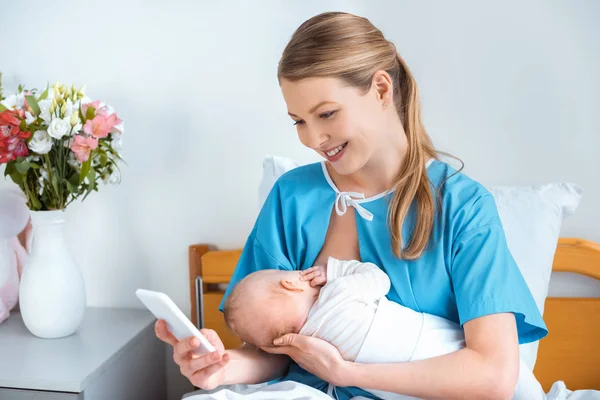 Beautiful smiling young mother breastfeeding baby and using smartphone in hospital room — Stock Photo