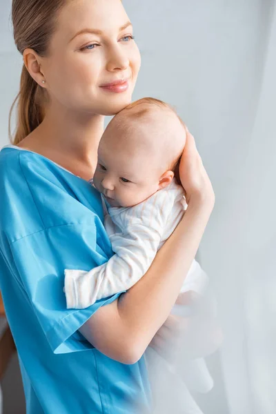 Schöne glückliche junge Mutter hält entzückendes Baby in der Hand und schaut im Krankenhauszimmer weg — Stockfoto