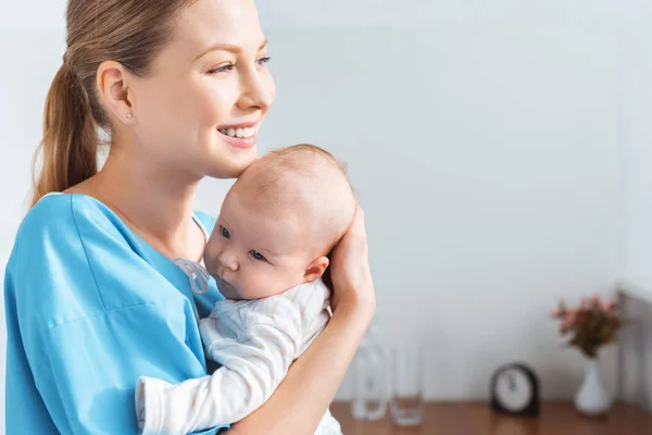 Side view of happy young mother holding adorable baby and looking away in hospital room — Stock Photo
