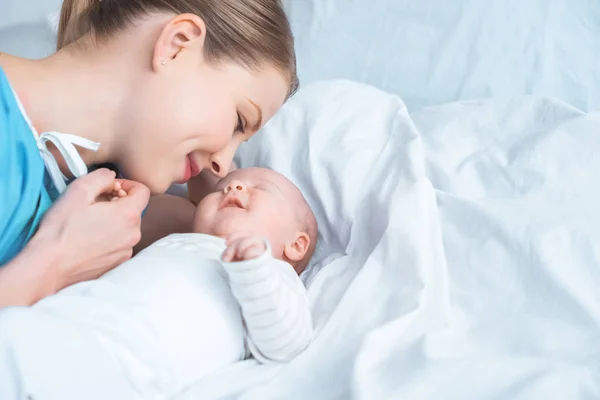 Smiling young mother holding hand of adorable newborn baby lying on bed — Stock Photo