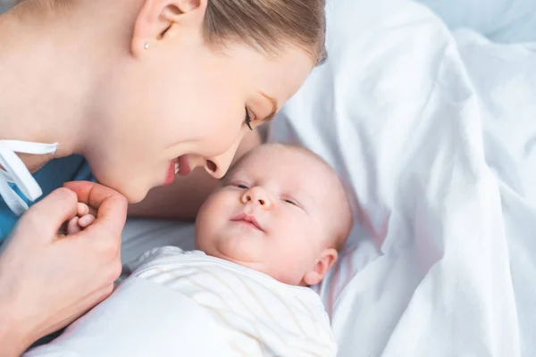 Счастливая молодая мать держит за руку очаровательного новорожденного ребенка, лежащего на кровати — стоковое фото