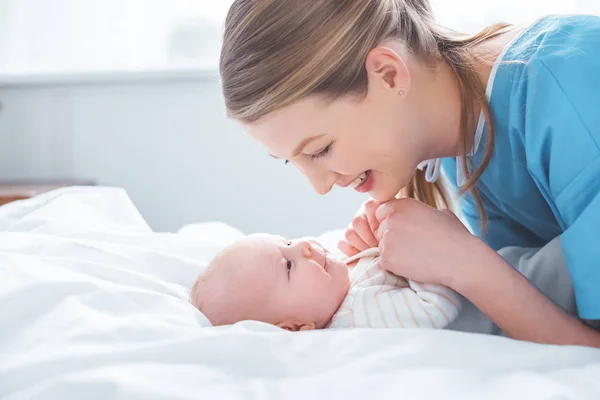 Vista lateral de la feliz madre joven cogida de la mano del adorable bebé en la habitación del hospital - foto de stock
