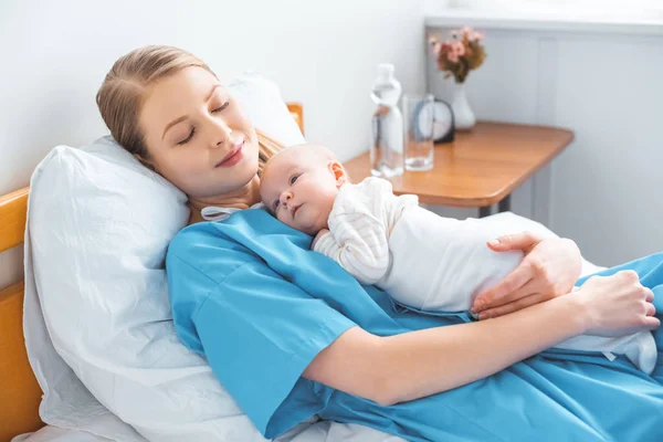 Young mother sleeping on hospital bed with adorable baby lying on chest — Stock Photo