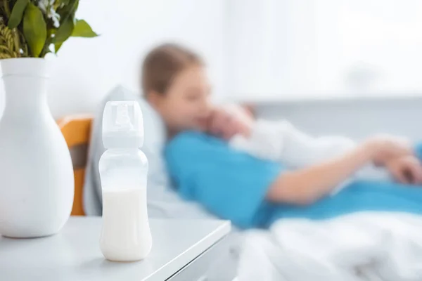 Close-up view of baby bottle with milk, vase and mother with newborn baby lying on hospital bed behind — Stock Photo