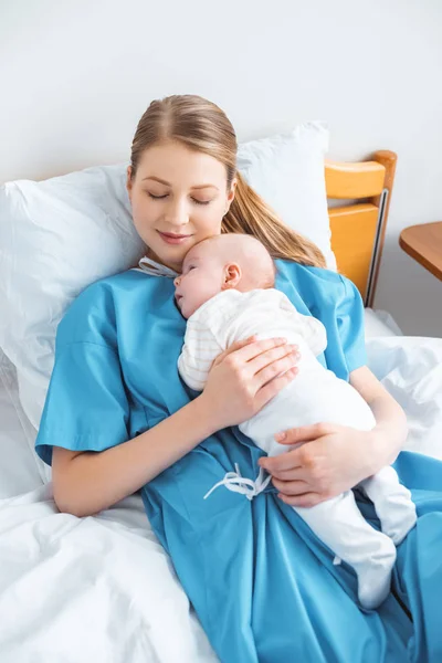 Feliz joven madre con los ojos cerrados acostado con adorable bebé en la habitación del hospital - foto de stock