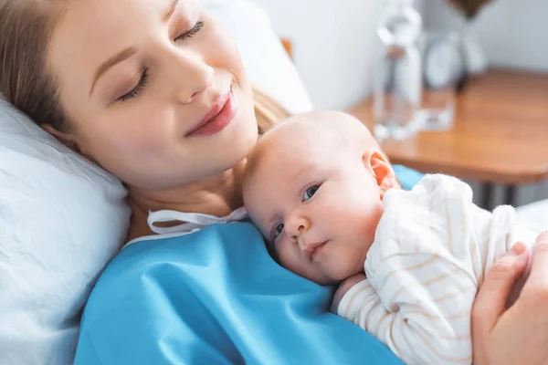 Happy young mother with closed eyes lying with adorable newborn baby on chest in hospital room — Stock Photo