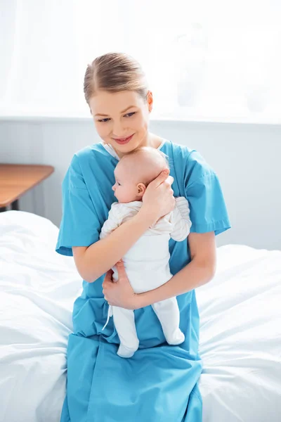 Високий кут зору щасливої молодої матері, що сидить на лікарняному ліжку і несе чарівну новонароджену дитину — стокове фото