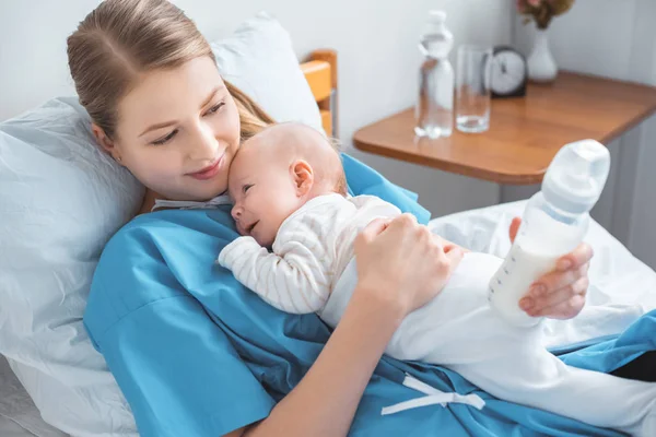 Vista de ángulo alto de sonriente madre joven sosteniendo biberón con leche y acostado en la cama con un bebé adorable - foto de stock