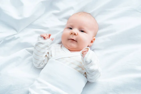 Top view of adorable baby looking at camera while lying on white bedding — Stock Photo