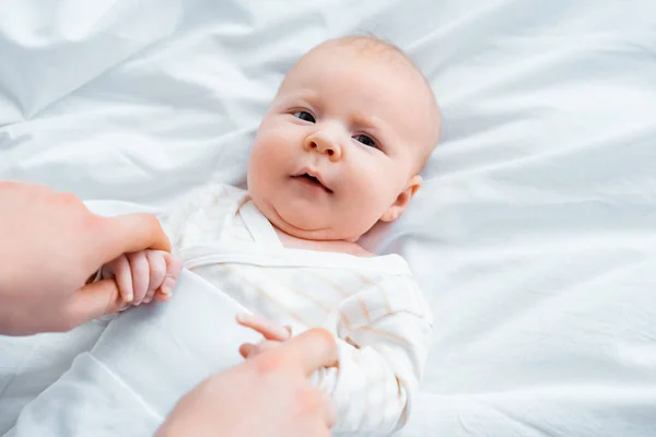 Abgeschnittene Aufnahme einer Mutter, die Hände eines entzückenden Säuglings hält, der auf weißem Bettzeug liegt — Stockfoto