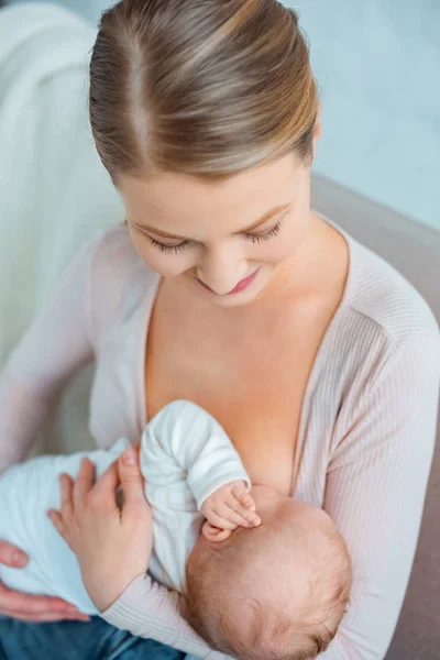 Vista de ángulo alto de hermosa mujer joven sonriente sentada en el sofá y amamantando a la hija lactante - foto de stock