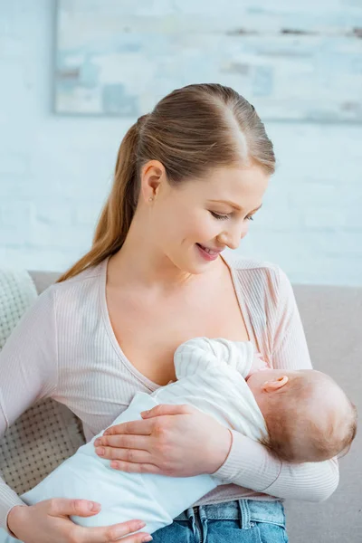 Hermosa sonriente joven sentada en el sofá y amamantando a su hija - foto de stock