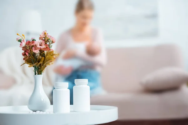 Recipientes com pílulas, flores em vaso e mãe bebê amamentando atrás em casa — Fotografia de Stock