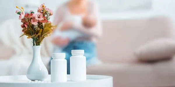 Vista close-up de recipientes com pílulas, flores em vaso e bebê de amamentação mãe atrás em casa — Fotografia de Stock