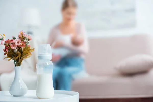 Close-up view of baby bottle with milk, flowers in vase and mother breastfeeding baby behind at home — Stock Photo