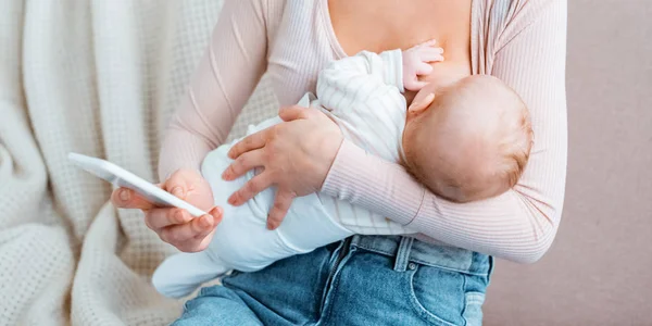 Inyección recortada de la madre joven amamantando al bebé y utilizando el teléfono inteligente - foto de stock
