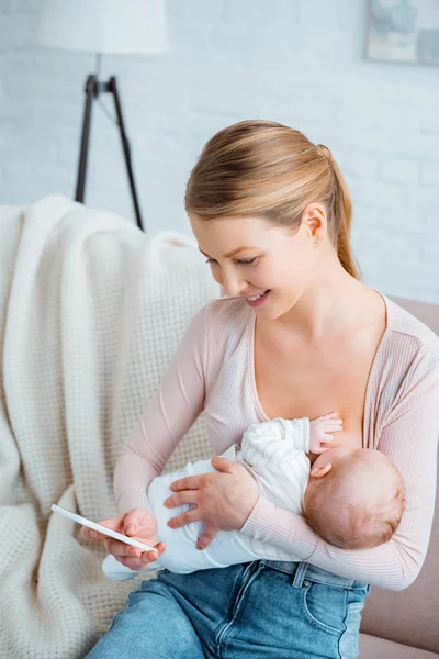 Vista de ángulo alto de la madre joven feliz amamantando al bebé y utilizando el teléfono inteligente en casa - foto de stock