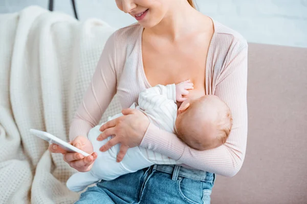 Recortado disparo de sonriente madre joven amamantando bebé y el uso de teléfono inteligente en casa - foto de stock