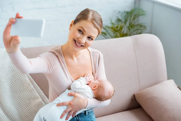 Jovem feliz amamentando bebê e tomando selfie com smartphone em casa — Fotografia de Stock