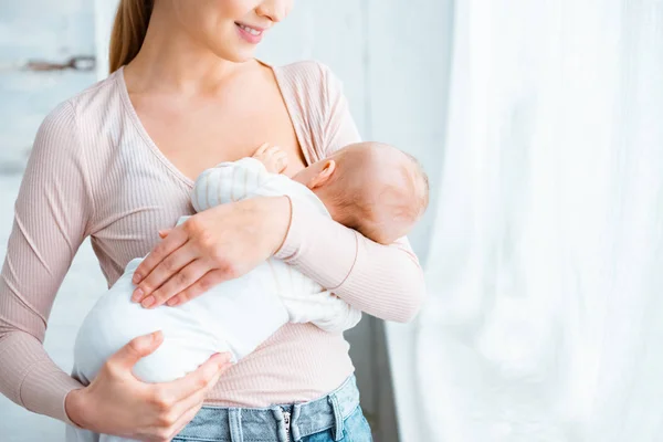 Обрезанный снимок улыбающейся молодой матери, стоящей и кормящей грудью младенца дома — стоковое фото