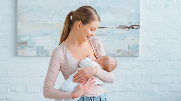 Schöne lächelnde junge Mutter, die ihr Baby zu Hause stillt — Stockfoto