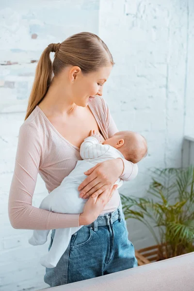 Schöne lächelnde junge Mutter, die ihr Baby zu Hause stillt — Stockfoto