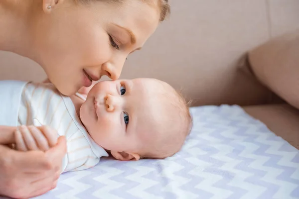 Cropped shot of happy mother kissing adorable infant child lying on couch and looking at camera — Stock Photo