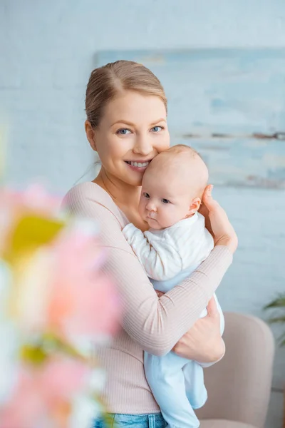 Enfoque selectivo de la madre joven y feliz abrazo adorable bebé y sonriendo a la cámara - foto de stock