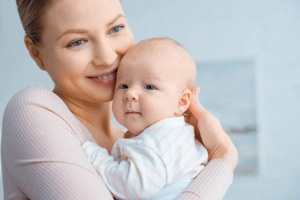 Feliz joven madre abrazando adorable bebé y mirando hacia otro lado - foto de stock