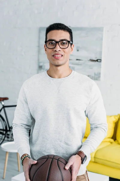 Handsome mixed race man in white sweatshirt and glasses holding brown ball — Stock Photo