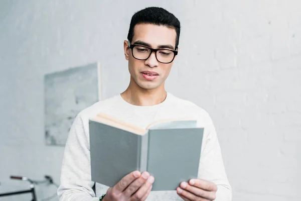 Jovem mestiço homem sério em camisola branca e óculos livro de leitura — Fotografia de Stock